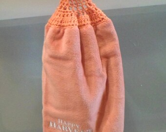 May include: A peach-colored kitchen towel with a crocheted loop at the top. The towel has a white embroidered border with the words "Happy Harvest" and a pattern of fall leaves.