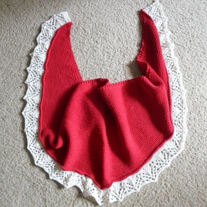 May include: A red knitted shawl with a white crocheted lace trim. The shawl is folded in half and laid flat on a light brown surface.