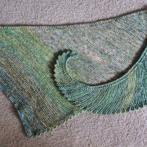 May include: A hand-knitted shawl in shades of green, blue, and yellow. The shawl has a triangular shape with a curved, scalloped edge. The yarn appears to have a subtle, variegated pattern, creating a textured look.
