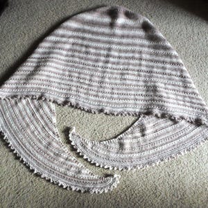 May include: A handmade, knitted shawl in shades of brown and white. The shawl features horizontal stripes and a scalloped edge. The triangular shape is designed to be worn over the shoulders. The soft, neutral colors make it a versatile accessory.