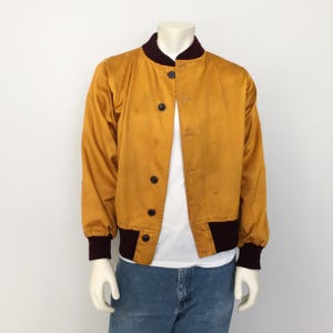 May include: Mustard yellow bomber jacket with a dark burgundy collar and cuffs. The jacket has a button-up front and black buttons. The jacket is displayed on a mannequin with blue jeans.