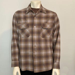 May include: A long-sleeve button-down shirt featuring a brown and light tan plaid pattern. The shirt includes a pointed collar, two chest pockets with flaps, and light-colored buttons running down the center front placket over a solid brown undershirt.
