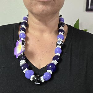 May include: A beaded necklace featuring a mix of purple, black, and white beads. The necklace includes a large purple flower and a black bead with a white floral design. The necklace is worn around the neck.