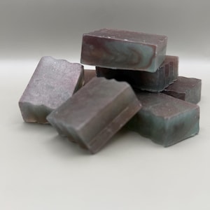 May include: A stack of rectangular soap bars featuring a marbled pattern of deep purple, dark grey, and muted teal tones. The bars have a smooth, solid texture with visible swirls of color throughout each piece, arranged in a small, clustered pile.