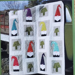 May include: A quilt featuring a repeating pattern of gnomes with colorful hats and beards. The quilt has a white background with gray squares. The gnomes have red, purple, blue, and yellow hats. The text "A Tale of Two Gnomes" is at the bottom.