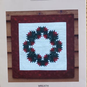 May include: A paper foundation pattern for a wreath, measuring 18.5 x 18.5 inches. The wreath design features a central white square with a wreath of dark green and red shapes. The pattern is from MH Designs.