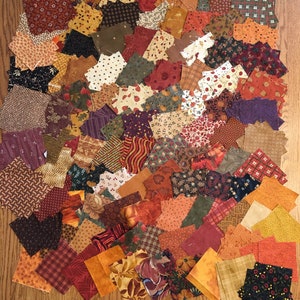 May include: A collection of fabric squares in various autumnal colors and patterns, including red, orange, brown, and green. The squares are arranged in a random pattern and are perfect for quilting or other craft projects.