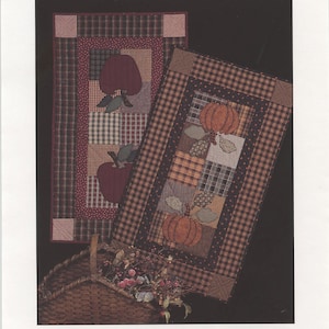 May include: Two quilted table toppers with a harvest theme. One features a red apple and the other a pumpkin. Both are made with a patchwork of brown, black, and white fabrics.