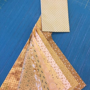May include: A collection of 15 fabric strips in various shades of yellow and brown with different patterns. The fabric strips are arranged in a fan shape on a blue surface. A small square piece of yellow fabric with black polka dots is also included.