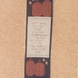 May include: A brown and black wall hanging with a recipe for pumpkin bars. The recipe is written in white on a white background. The wall hanging is 10 inches by 31 inches and features two brown pumpkins with a black background. The text "Farmyard Creations" is at the bottom of the image.