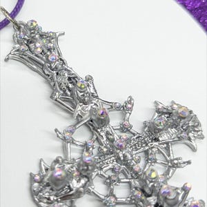 May include: Silver cross pendant with iridescent rhinestones. The cross has an intricate, openwork design. A purple cord is attached. The pendant is approximately 7.6 cm long.