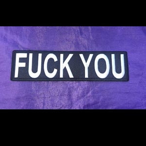 May include: A rectangular black patch with the words "FUCK YOU" in bold, white capital letters. The patch is set against a purple background.