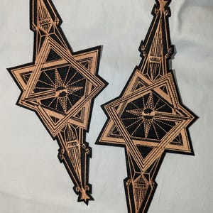 May include: Two black and gold embroidered patches with a starburst design. The patches are shaped like a star with a pointed top and bottom.