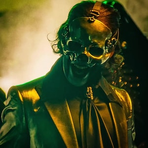 May include: A person wearing a black suit jacket with a gold skull mask. The mask has a leather strap and is covering the person's face. The person is standing in front of a green and yellow background.
