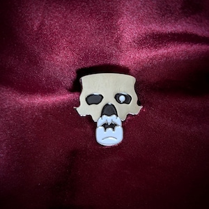 Könnte beinhalten: Eine silberfarbene, schädelformige Anstecknadel mit schwarzen und weißen Emaille-Details. Die Anstecknadel zeigt ein stilisiertes Schädeldesign mit einem offenen und einem geschlossenen Auge. Die Anstecknadel befindet sich auf einem tiefroten Hintergrund.