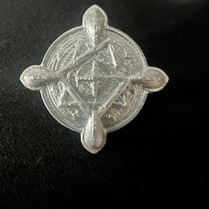 May include: A silver-colored, circular pendant with a detailed, symmetrical design. The pendant features a cross-like shape with ornate detailing and four teardrop-shaped extensions. The intricate pattern suggests a Celtic or historical theme.