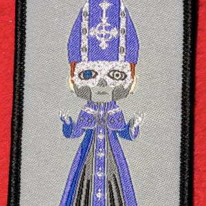 Puede incluir: Parche bordado con las palabras "Blasfemia" y "Herejía". El parche presenta una figura de estilo caricatura con un atuendo papal azul y plateado con una cara de calavera. El parche tiene un borde negro y está sobre un fondo gris.