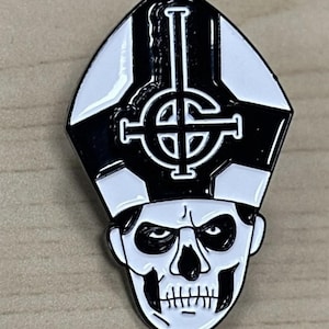 May include: A black and white enamel pin featuring a skull-like face wearing a bishop's mitre. The pin has a detailed design with a cross-like symbol on the mitre. The skull has a stern expression with visible teeth.