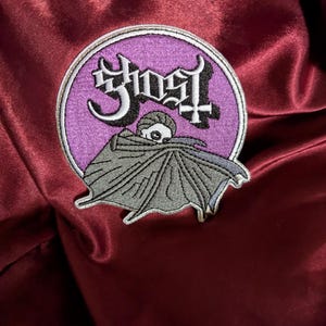 May include: Embroidered patch with the band name "Ghost" in a stylized font, set within a purple and white circular design. The patch features a gray cloaked figure. The patch is set against a burgundy satin background.