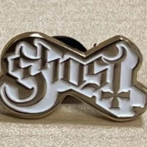 Könnte beinhalten: Goldfarbene Metallnadel mit dem Wort "GHOST" in weißer, stilisierter Schrift. Die Nadel hat ein dekoratives Design und einen Nadelverschluss.