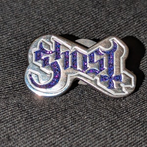 Peut inclure: Épingle en métal argenté avec le mot "Ghost" dans une police stylisée, rempli de paillettes violettes. L'épingle est posée sur un fond gris foncé texturé.