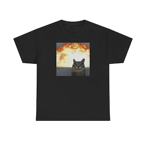 May include: Black t-shirt featuring a meme of a cat with a nuclear explosion in the background. The cat is gray and brown with green eyes. The explosion is orange and yellow.
