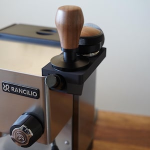 Puede incluir: Máquina de espresso de acero inoxidable con un tamper negro y de madera y un embudo dosificador negro. El tamper tiene un mango de madera y una base negra. El logotipo "Rancilio" es visible en la máquina.