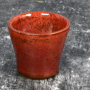 May include: A red ceramic cup with a speckled glaze. The cup is glazed inside and out and has a rounded bottom.