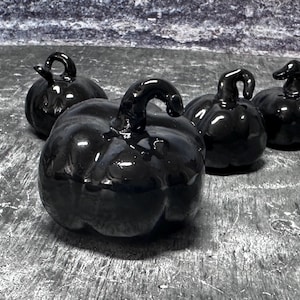 May include: Four glossy black ceramic pumpkins with curved stems are arranged on a textured surface. The pumpkins vary slightly in size and stem shape, creating a decorative display. The pumpkins are ideal for fall or Halloween decor.