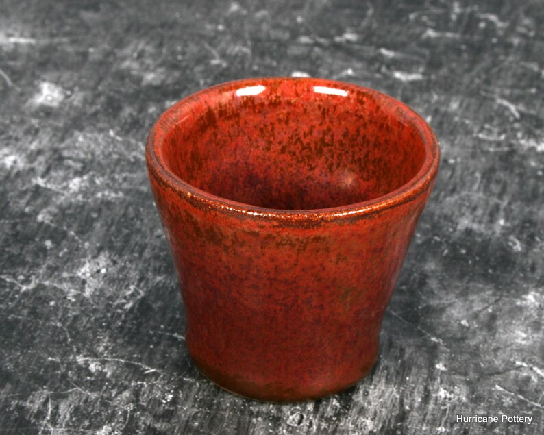 This is a single shot glass glazed in dark orange-red  glaze with speckles and crystals of red-brown, giving a copper appearance. The surface is smooth with little to no texture.