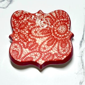 May include: A decorative, red ceramic dish with a white lace pattern. The dish has a unique, ornate shape with curved edges. The lace design is detailed and covers the entire surface of the dish. The dish is likely used for serving or display.
