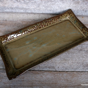 May include: A rectangular ceramic serving tray with a brown and tan glaze. The tray has a slightly curved shape with a textured brown rim. The interior has subtle blue accents. The tray is handmade and suitable for serving food.