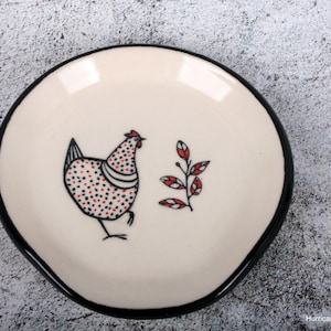 May include: Cream-colored ceramic spoon rest with a black rim. The center features a whimsical illustration of a chicken with red and black polka dots, and a sprig of red leaves. A functional and decorative kitchen accessory.