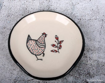 May include: Cream-colored ceramic spoon rest with a black rim. The center features a whimsical illustration of a chicken with red and black polka dots, and a sprig of red leaves. A functional and decorative kitchen accessory.