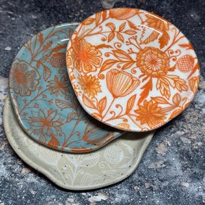 May include: Three ceramic dishes with floral patterns. The top dish is white with orange flowers, the middle dish is light blue with orange flowers, and the bottom dish is beige with white flowers. The dishes are stacked on top of each other.