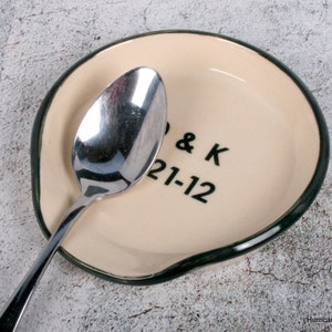 May include: A white ceramic spoon rest with a green rim. The spoon rest has the text "J & K 21-12" written on it in green.