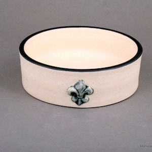 May include: A white ceramic bowl with a black rim and a green fleur-de-lis design. The bowl is perfect for serving snacks or dips.