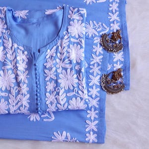 Puede incluir: Un conjunto azul claro con una túnica bordada y tela a juego, junto con un par de pendientes plateados ornamentados. La túnica presenta bordados florales blancos y una botonadura. Los pendientes tienen un diseño detallado.