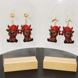 May include: Christmas earrings featuring reindeer figures with red hats and scarves. The brown reindeer have black accents, and the earrings have gold-coloured snowflake or bow details. The earrings are displayed on clear acrylic stands with wooden bases.