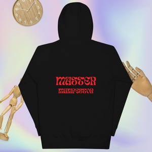 May include: Black hooded sweatshirt with the words "MASTER MANIFESTOR" in red, stylized lettering. The back of the sweatshirt is the focus of the image. A wooden clock and a wooden hand are in the background.