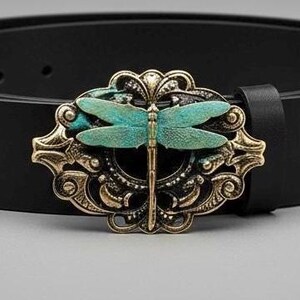 May include: A black belt featuring a decorative oval buckle with an ornate gold-toned metal frame and a central turquoise-colored dragonfly motif. The belt strap is smooth and solid black, secured by a matching black leather loop near the buckle.