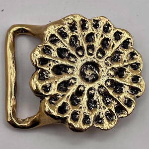 May include: A gold-toned metal belt buckle featuring a circular, flower-like design with a textured, dark recessed center and a smooth, raised outer edge. The buckle has a rectangular loop on the left side for attaching to a belt strap.