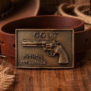 May include: A brown leather belt with a rectangular brass buckle. The buckle features the word "COLT" above a detailed image of a revolver, with "PYTHON .357 MAGNUM" below. The belt is on a wooden surface.