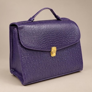 May include: A deep purple handbag featuring a textured ostrich-skin pattern on the body and a crocodile-embossed flap. The bag includes a single top handle and a central gold-toned metal push-lock closure on the front flap.