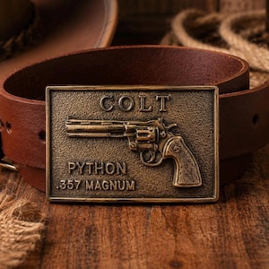 May include: A brown leather belt featuring a rectangular brass-toned metal buckle. The buckle displays a raised relief of a revolver with the text COLT, PYTHON, and .357 MAGNUM embossed on the surface in a clear, capitalized font style.