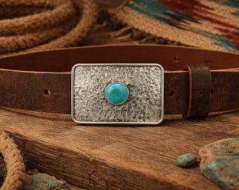May include: A brown leather belt featuring a rectangular metal buckle with a hammered, textured finish and a single round, polished turquoise stone set in the center. The belt includes a matching leather keeper loop and pre-punched holes.