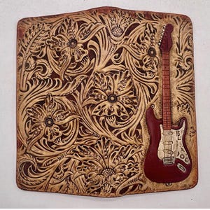 May include: A rectangular leather piece with a carved floral design and a burgundy electric guitar. The guitar features a white pickguard and the initials "BL". The background is light brown. The piece is likely hand-tooled.