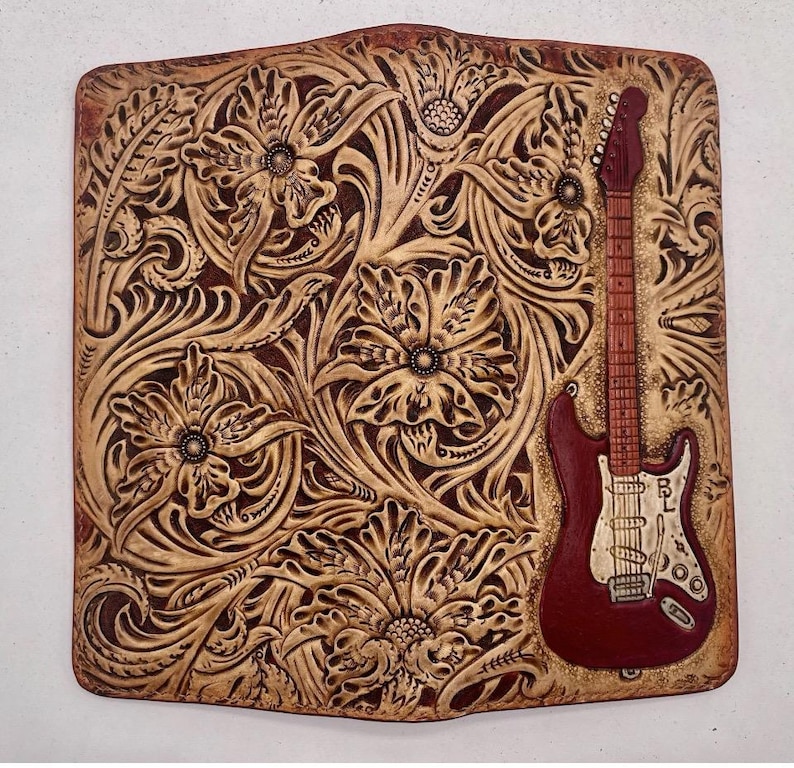 May include: A rectangular leather piece with a carved floral design and a burgundy electric guitar. The guitar features a white pickguard and the initials "BL". The background is light brown. The piece is likely hand-tooled.