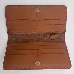 May include: Open, tan leather wallet with multiple card slots, a zippered compartment, and snap closures. The wallet is rectangular and features a textured interior lining. The hardware is a brass colour.