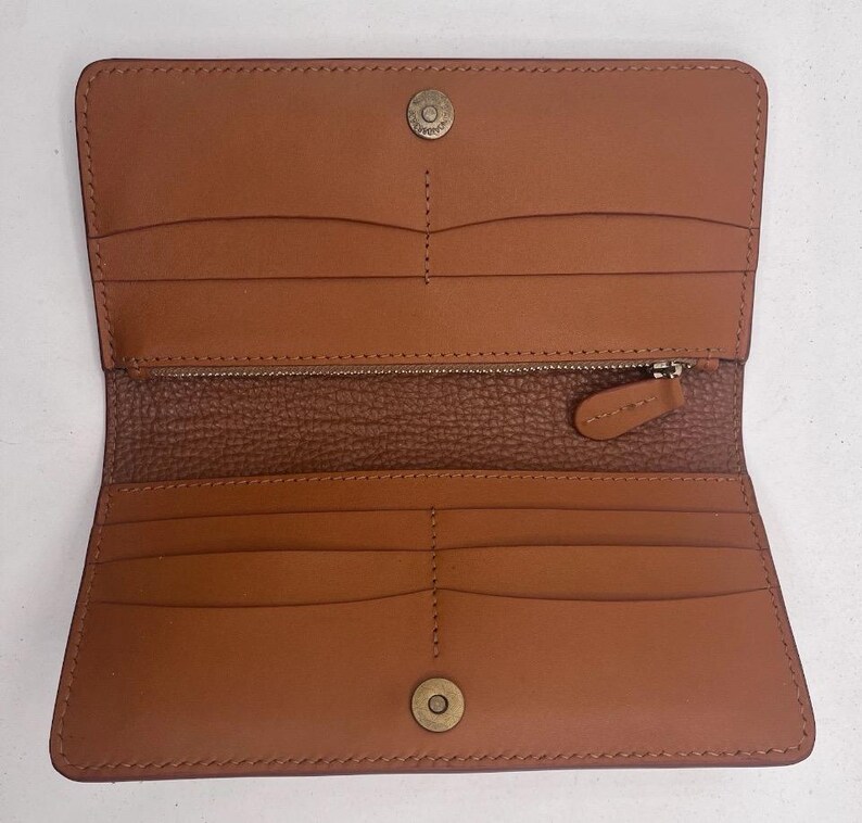May include: Open, tan leather wallet with multiple card slots, a zippered compartment, and snap closures. The wallet is rectangular and features a textured interior lining. The hardware is a brass colour.